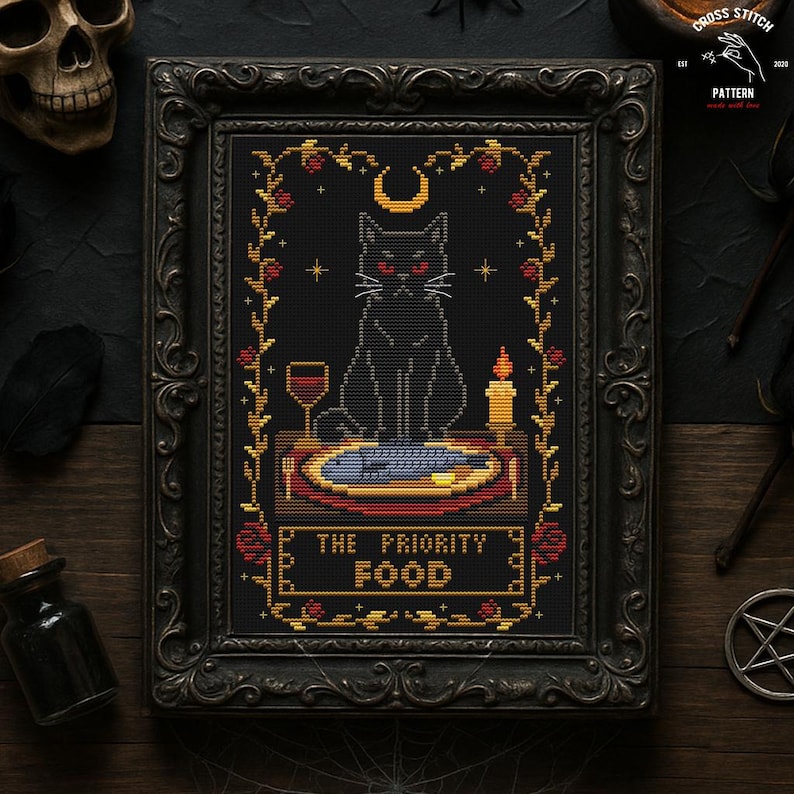 May include: Cross-stitch art in a black ornate frame. The design features a black cat with red eyes, a crescent moon, and the text "THE PRIORITY FOOD." A glass of wine, a candle, and a fish on a plate are also depicted.