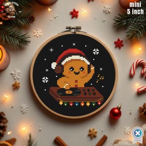 May include: A 5-inch (12.7 cm) diameter cross-stitch hoop featuring a gingerbread man wearing a Santa hat and headphones. The gingerbread man is on a brown platform with colorful lights. The background is black with white snowflakes.