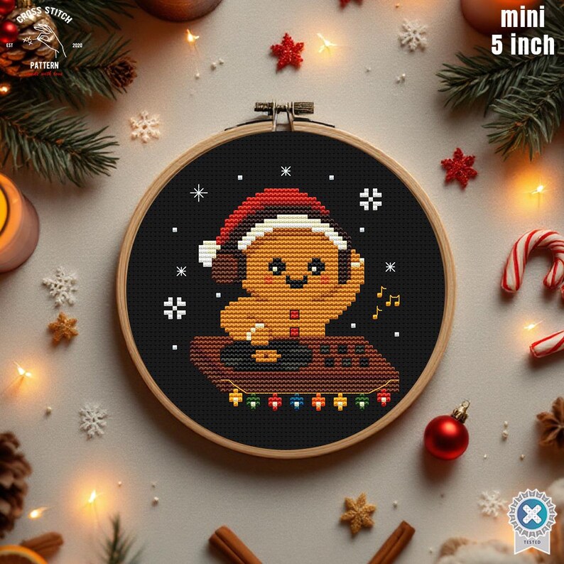 May include: A 5-inch (12.7 cm) diameter cross-stitch hoop featuring a gingerbread man wearing a Santa hat and headphones. The gingerbread man is on a brown platform with colorful lights. The background is black with white snowflakes.