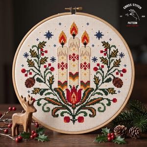 May include: A round wooden embroidery hoop containing a cross stitch pattern of three lit candles, holly leaves, red berries, and pinecones. The design features a central red flower motif and small blue stars. Text in the corner reads CROSS STITCH PATTERN EST 2020 made with love.