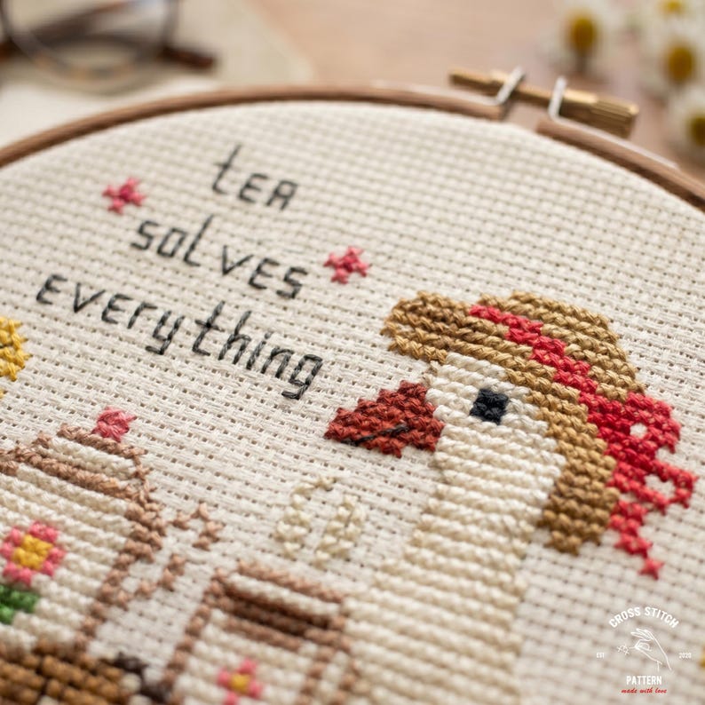 May include: Cross-stitch embroidery in a wooden hoop with the text "Tea Solves Everything." The design features a goose with a red bow, a teapot, and flowers, all stitched in various colors on a beige fabric.