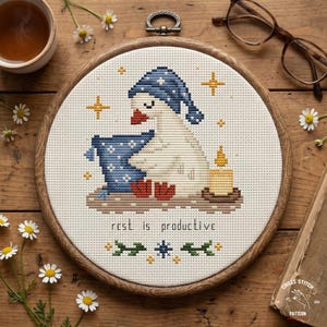 May include: Cross-stitch embroidery in a wooden hoop features a goose wearing a blue nightcap and holding a pillow. A lit candle and the text "rest is productive" are also included. The design is surrounded by gold stars and floral accents.