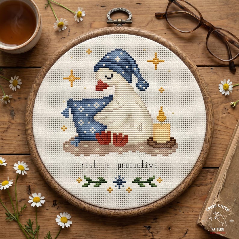 May include: Cross-stitch embroidery in a wooden hoop features a goose wearing a blue nightcap and holding a pillow. A lit candle and the text "rest is productive" are also included. The design is surrounded by gold stars and floral accents.