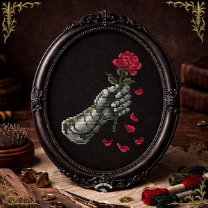 May include: Cross-stitch art in a black oval frame. The artwork depicts a skeletal hand holding a red rose with falling petals. The frame has ornate detailing. The background is black.