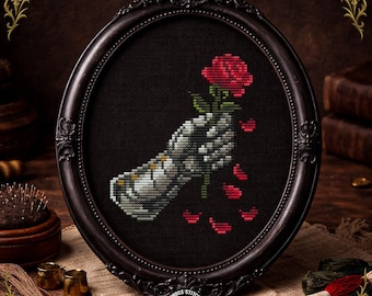 Pode incluir: Arte de ponto cruz em uma moldura oval preta. A obra de arte retrata uma mão esquelética segurando uma rosa vermelha com pétalas caindo. A moldura tem detalhes ornamentados. O fundo é preto.