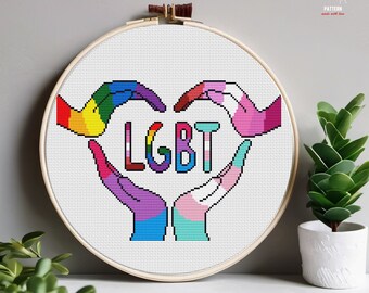 May include: Cross stitch pattern featuring a heart formed by two hands, each with a different pride flag color scheme. The word "LGBT" is stitched in the center of the heart.