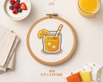 Pode incluir: Um bastidor de bordado em ponto cruz com um desenho de cocktail pixelizado, com uma fatia de laranja e uma palhinha. O bastidor tem 6,88 x 8,36 cm. A imagem inclui rolos de linha, um copo de sumo de laranja e uma taça de iogurte com frutos silvestres.