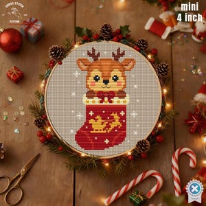 Może przedstawiać: Mini wzór haftu krzyżykowego o wymiarach 10 cm przedstawiający kreskówkowego renifera w czerwonej świątecznej skarpecie. Haft umieszczony jest w drewnianym tamborku, otoczonym szyszkami, jagodami i światełkami. Projekt zawiera płatki śniegu i złotego renifera.