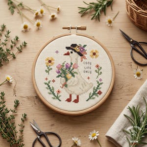 Peut inclure: Un cercle de broderie au point de croix avec un motif d'oie portant un chapeau et tenant des fleurs. Le motif comprend le texte "Cozy Life Only" et est entouré d'éléments floraux. Le cercle est posé sur une surface en bois avec des ciseaux et des brins de verdure.