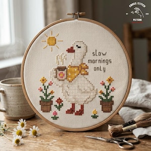 May include: Cross-stitch embroidery in a wooden hoop features a goose holding a coffee cup, wearing a yellow scarf, and the text "slow mornings only." The design includes a sun, flowers, and the words "Cross Stitch Pattern."