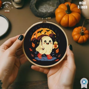 May include: A mini 5-inch cross-stitch embroidery hoop featuring a cute ghost under an umbrella, holding a pumpkin spice latte. The design includes autumn leaves and rain on a black background. The text "Cross Stitch Pattern" is visible.