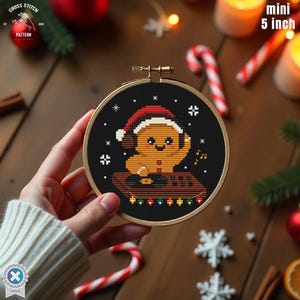 May include: A mini 5-inch cross-stitch embroidery hoop featuring a gingerbread man wearing a Santa hat and headphones. The design includes musical notes and decorative lights on a black background, perfect for holiday decor.