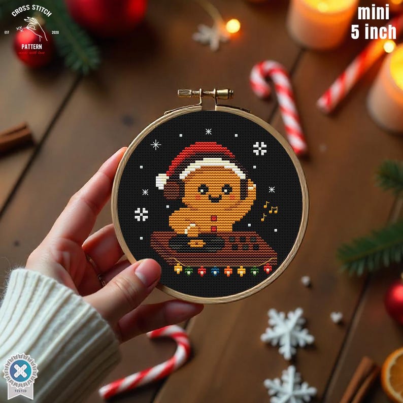 May include: A mini 5-inch cross-stitch embroidery hoop featuring a gingerbread man wearing a Santa hat and headphones. The design includes musical notes and decorative lights on a black background, perfect for holiday decor.