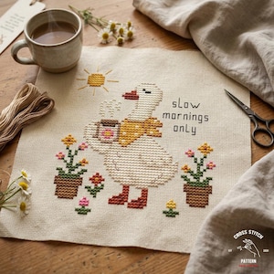 May include: Cross-stitch pattern featuring a goose holding a coffee cup, with the text "slow mornings only." The design includes potted flowers, a sun, and a yellow scarf. The background is a beige fabric.
