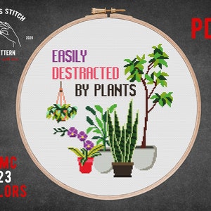 以下が含まれることがあります： 「Easily Distracted by Plants」というテキストと、吊り下げられた植物、ヘビの植物、鉢植えの木など、さまざまな鉢植えの植物が描かれたクロスステッチパターン。パターンは、木製のフレームが付いた白い刺繍枠に入っています。画像には「DMC 23 Colors」というテキストも含まれています。