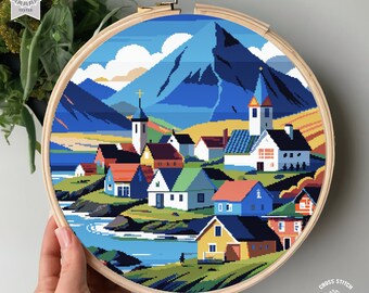 May include: A cross stitch pattern featuring a colorful village nestled in a mountain valley. The scene includes a church, houses, a river, and a mountain range in the background. The pattern is framed in a wooden hoop.