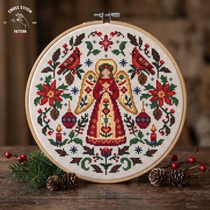 May include: A circular wooden embroidery hoop containing a cross stitch pattern of a central angel figure in red and gold robes, flanked by red cardinals, poinsettias, pinecones, and candles on a white fabric background with a festive design.