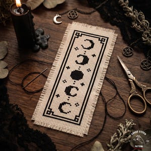 Può includere: Un segnalibro beige a punto croce con un disegno nero di fasi lunari e stelle. Il segnalibro ha un bordo sfilacciato. Forniture per cucito, una candela e ciondoli circondano il segnalibro. Il testo "CROSS STITCH PATTERN" è incluso.