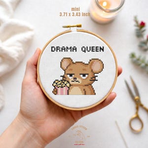 Może przedstawiać: Mini obręcz do haftu krzyżykowego z napisem "DRAMA QUEEN" nad kreskówkową myszą trzymającą popcorn. Obręcz ma wymiary 9,42 x 8,71 cm. Mysz ma zrzędliwą minę.