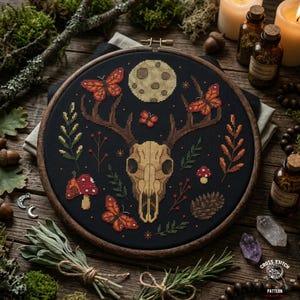 May include: A cross-stitch embroidery hoop featuring a deer skull, butterflies, and a full moon against a black background. The design includes mushrooms, leaves, and berries in shades of orange, green, and brown. The hoop is framed in wood.
