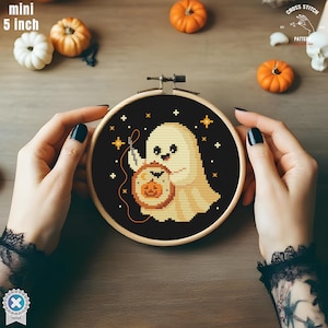 May include: A cross-stitch embroidery hoop featuring a Halloween ghost holding a pumpkin lantern. The design is set against a black background with stars. The hoop is 12.7 cm in diameter. The text "mini 5 inch" is visible.