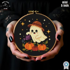 以下が含まれることがあります： ハロウィーンのデザインが施された、直径12.7cmのミニクロスステッチ刺繍枠。茶色の帽子をかぶったお化けが紫色のじょうろでカボチャに水をやり、黒い背景に星が描かれています。「CROSS STITCH PATTERN」の文字が見えます。