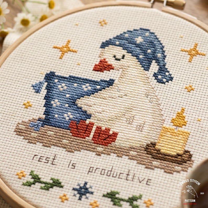 May include: Cross-stitch embroidery of a goose wearing a blue nightcap and resting on a pillow. The goose has red feet and a candle is lit nearby. The text "rest is productive" is stitched below the goose.