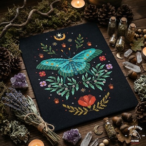 May include: A cross-stitch pattern featuring a teal butterfly with intricate details, surrounded by floral and celestial elements on a black background. The design includes flowers, leaves, and a crescent moon. The image is set on a wooden surface with candles and crystals.
