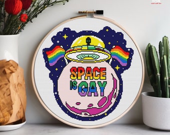 May include: A cross stitch pattern featuring a pink planet with the text "SPACE IS GAY" in rainbow colors. A rainbow flag and a UFO with a rainbow flag are above the planet. The pattern is on a white background.