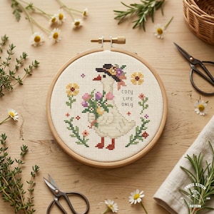 May include: A cross-stitch embroidery hoop featuring a goose wearing a hat and holding flowers. The design includes the text "COZY LIFE ONLY" and is surrounded by floral elements. The hoop is set on a wooden surface with scissors and sprigs of herbs.