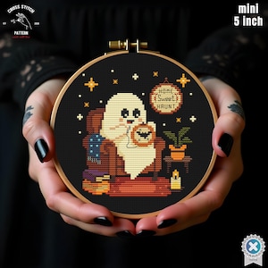 Könnte beinhalten: Ein 12,7 cm Mini-Kreuzstich-Stickrahmen mit einem Halloween-Motiv. Das Design zeigt einen Geist, der einen Kürbis hält, ein Schild mit der Aufschrift "Home Sweet Haunt" und andere dekorative Elemente. Der Rahmen wird in den Händen einer Person gehalten.