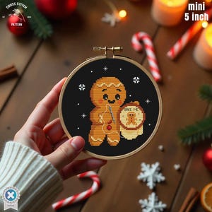 Può includere: Un mini telaio da ricamo a punto croce da 12,7 cm con un omino di pan di zenzero che tiene un barattolo con la scritta "BAKE ME". Il disegno è ambientato su uno sfondo nero con accenti di fiocchi di neve bianchi. Il telaio è tenuto in mano.