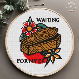 May include: Cross-stitch embroidery in a wooden hoop. The design features a coffin, flowers, and the text "WAITING FOR MY EX" in black. The coffin is brown and black, set against a red background.