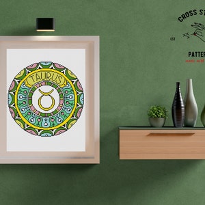 Peut inclure: Un motif de point de croix encadr&eacute; du signe du zodiaque Taureau. Le motif repr&eacute;sente une t&ecirc;te de taureau entour&eacute;e d'un mandala color&eacute;. Le cadre est blanc avec une finition en bois clair. Le texte "CROSS STITCH PATTERN made with love" est visible dans le coin sup&eacute;rieur droit.