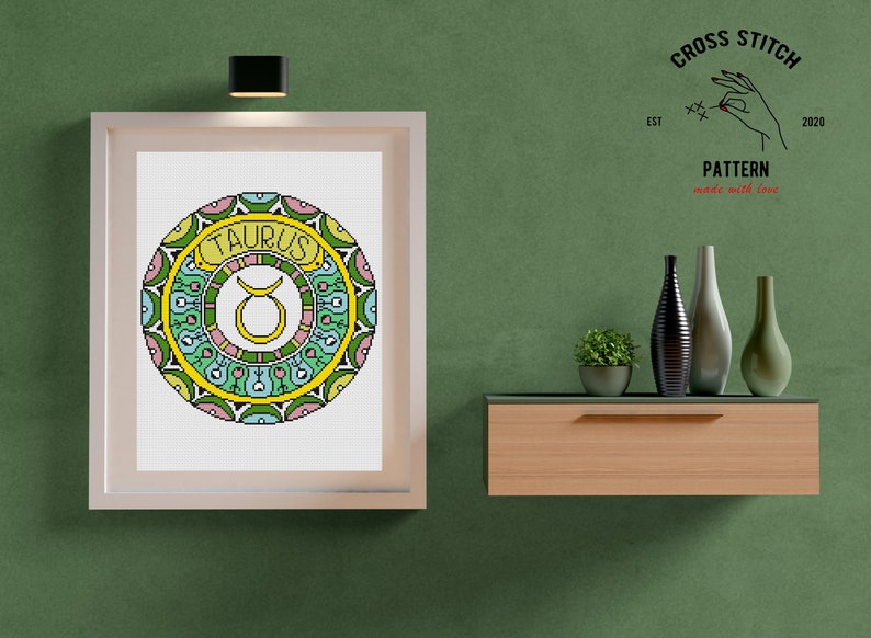 Peut inclure: Un motif de point de croix encadr&eacute; du signe du zodiaque Taureau. Le motif repr&eacute;sente une t&ecirc;te de taureau entour&eacute;e d'un mandala color&eacute;. Le cadre est blanc avec une finition en bois clair. Le texte "CROSS STITCH PATTERN made with love" est visible dans le coin sup&eacute;rieur droit.