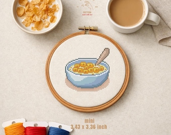 Pode incluir: Um bastidor de bordado em ponto cruz com um desenho de tigela de cereais com uma colher. A imagem inclui uma tigela de cereais com leite, uma chávena de café e bobinas de linha. O texto diz "CROSS STITCH PATTERN made with love" e "mini 8.71cm x 8.53cm".