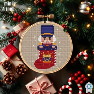 Könnte beinhalten: Ein Mini-Kreuzstichmuster von 10 cm in einem Holzreifen. Das Design zeigt einen Nussknackersoldaten in einem roten Strumpf mit einem goldenen Emblem. Der Hintergrund ist ein neutraler Beigeton mit weißen Schneeflocken. Der Text "CROSS STITCH PATTERN" ist sichtbar.