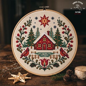 May include: A circular wooden embroidery hoop containing a cross stitch pattern of a red cabin in a snowy forest. The design features two red cardinals, green pine trees, a large star, and floral motifs in red, green, and white on cream fabric.