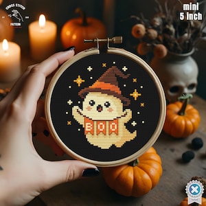 Könnte beinhalten: Ein Mini-Stickrahmen von 12,7 cm mit einem Halloween-Geist, der einen Hexenhut und ein Banner mit der Aufschrift "BOO" trägt. Der Geist ist von Sternen auf schwarzem Hintergrund umgeben. Der Stickrahmen wird in einer Hand gehalten.