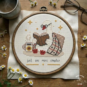 May include: Cross-stitch embroidery in a wooden hoop features a swan reading a book, a cup of coffee, and the text "just one more chapter." The design includes yellow stars and red hearts. The embroidery is on a cream-colored fabric.