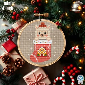 May include: A cross-stitch embroidery hoop featuring a festive design. The artwork depicts a cute llama wearing a Santa hat, peeking out of a Christmas stocking. The stocking has a house design. The hoop is 4 inches (10 cm) in diameter.