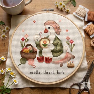 May include: Cross-stitch embroidery in a wooden hoop featuring a goose wearing a hat and vest, holding a needle and thread. The design includes a basket of fruit, flowers, and the text "needle, thread, honk".