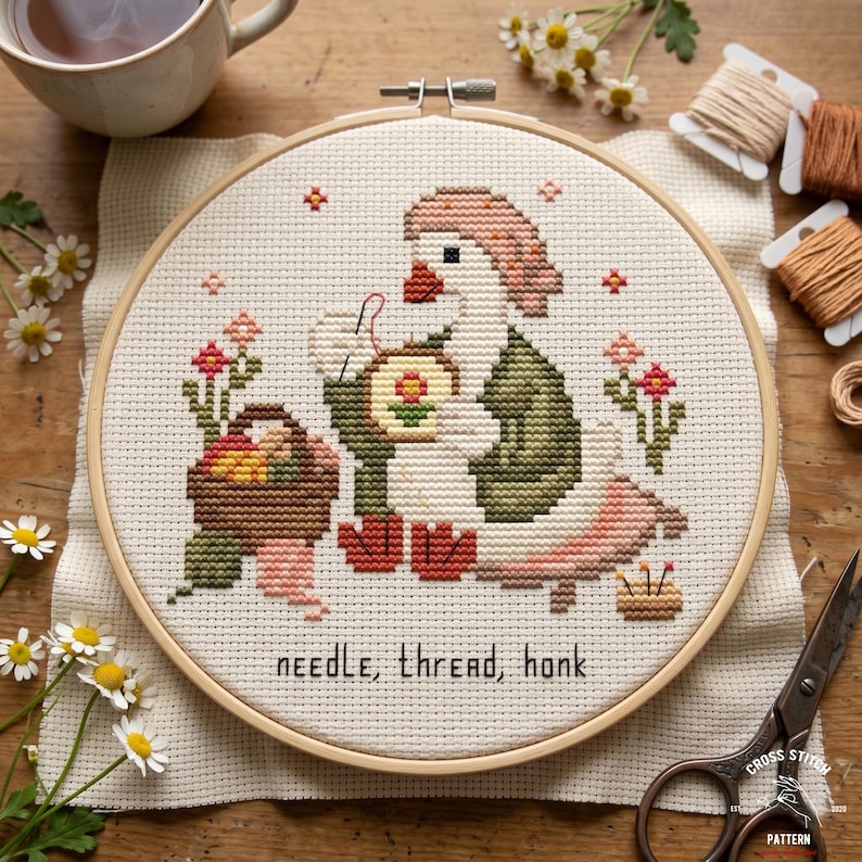 May include: Cross-stitch embroidery in a wooden hoop featuring a goose wearing a hat and vest, holding a needle and thread. The design includes a basket of fruit, flowers, and the text "needle, thread, honk".