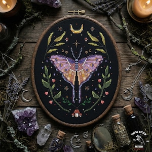 Puede incluir: Un patrón de punto de cruz con una polilla de alas moradas y naranjas, rodeada de hojas verdes, una luna creciente y estrellas. El diseño está sobre un fondo negro dentro de un bastidor de bordado de madera.