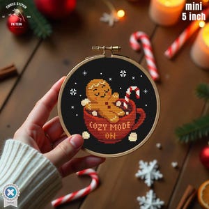 Peut inclure: Un mini cercle à broder de 12,7 cm avec un bonhomme en pain d'épices dans une tasse rouge. Le motif comprend le texte "COZY MODE ON", des flocons de neige et une canne en sucre sur fond noir.