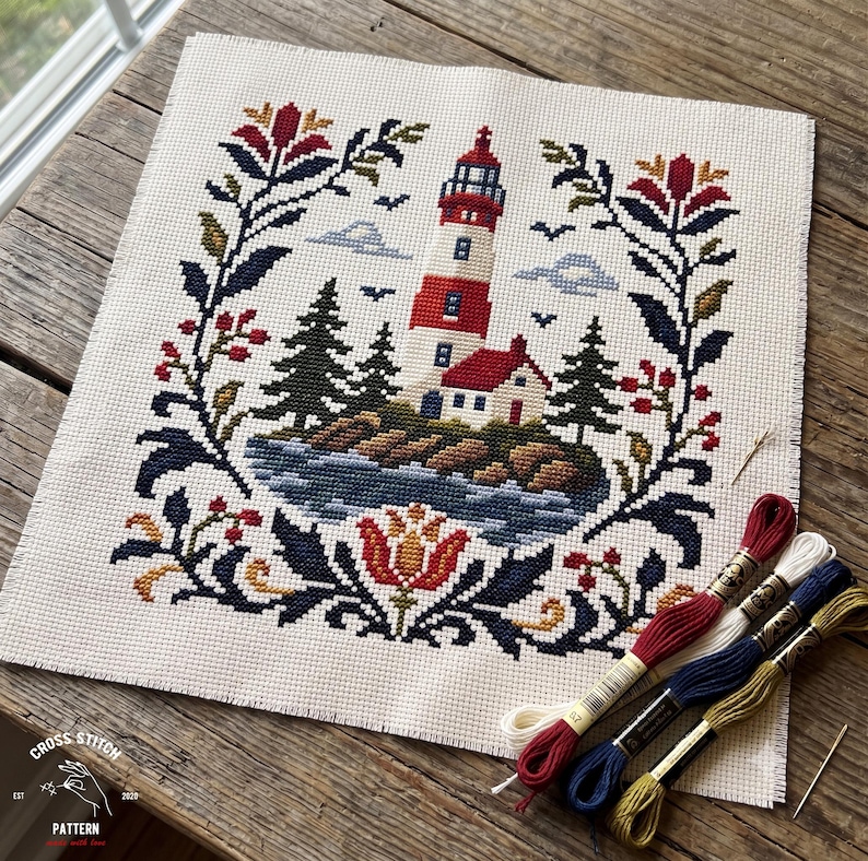 May include: A cross-stitch pattern on cream fabric featuring a red and white lighthouse on a rocky island with pine trees, surrounded by a floral border in red, blue, and gold. Four skeins of embroidery floss in red, white, blue, and gold are included.