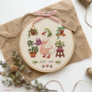 May include: A cross-stitch hoop art piece featuring a goose watering plants. The design includes the words "plant mom" and a tag that says "for plant lovers". The hoop is tied with a pink ribbon.