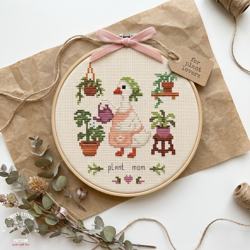 May include: A cross-stitch hoop art piece featuring a goose watering plants. The design includes the words "plant mom" and a tag that says "for plant lovers". The hoop is tied with a pink ribbon.