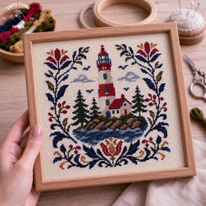 Peut inclure: Une broderie au point de croix carrée dans un cadre en bois clair représentant un phare rouge et blanc sur une île rocheuse avec des pins, entourée d'une bordure florale symétrique en fil bleu marine, rouge et doré sur tissu crème.