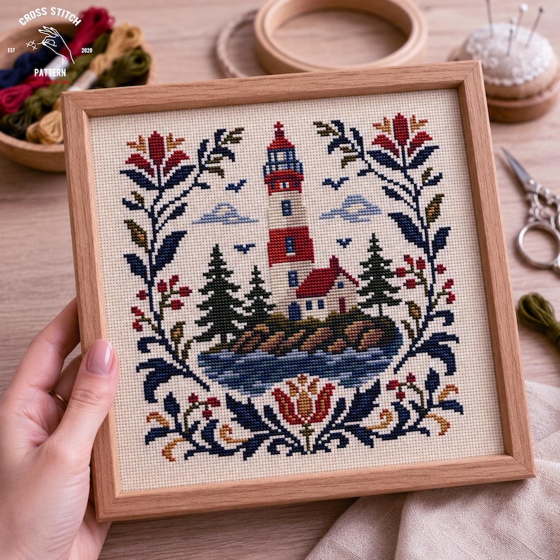 May include: A square cross stitch embroidery in a light wood frame featuring a red and white lighthouse on a rocky island with pine trees, surrounded by a symmetrical floral border in navy blue, red, and gold thread on cream fabric.